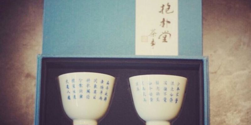 A pair of white porcelain cups with blue calligraphy, displayed open inside a teal gift box