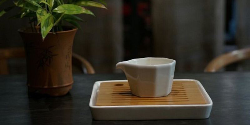 A white octagonal porcelain pitcher resting on a wooden tray, beside a potted plant