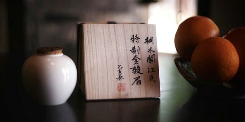 A small white ceramic jar beside a wooden box with calligraphy, with oranges blurred in the background