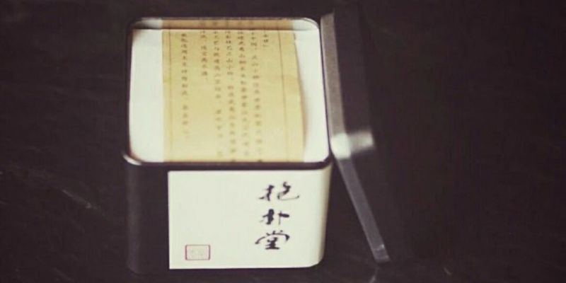 一只打开的方形茶罐，露出印有文字的内衬纸，罐盖斜靠在罐身旁