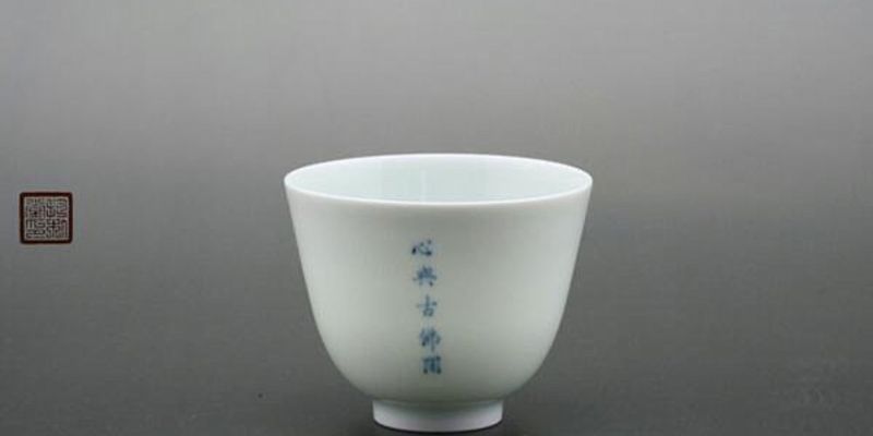 A single pale porcelain cup with vertical blue calligraphy, photographed against a grey studio background