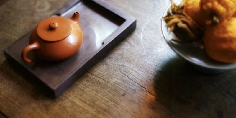 A small zisha teapot resting on a dark rectangular wooden tray, beside a bowl of citrus fruit
