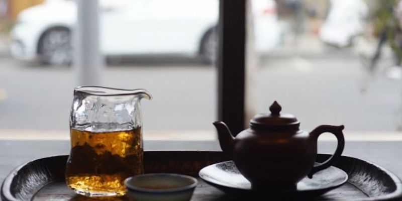 玻璃公道杯中盛着琥珀色茶汤，旁边是一只深色小茶壶，背景透过窗户可见模糊的街景