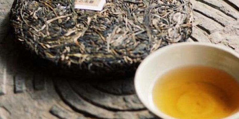 一饼普洱生茶饼置于一杯金黄茶汤旁，背景是带雕刻纹路的石面