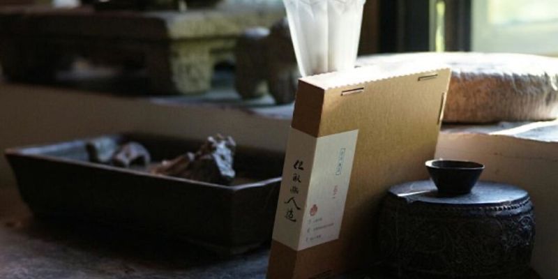 光线昏暗的茶室一角，石桌上摆着一盒包装好的茶，旁边是黑陶罐与茶杯