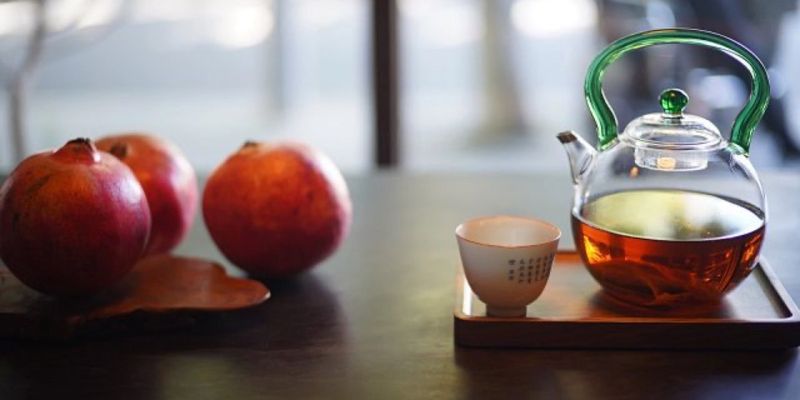 A glass teapot of brewed amber tea beside a small ceramic cup on a wooden tray, with three pomegranates in the background