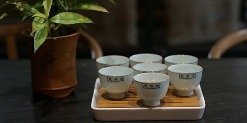Seven small white porcelain tea cups arranged on a wooden tray beside a potted plant