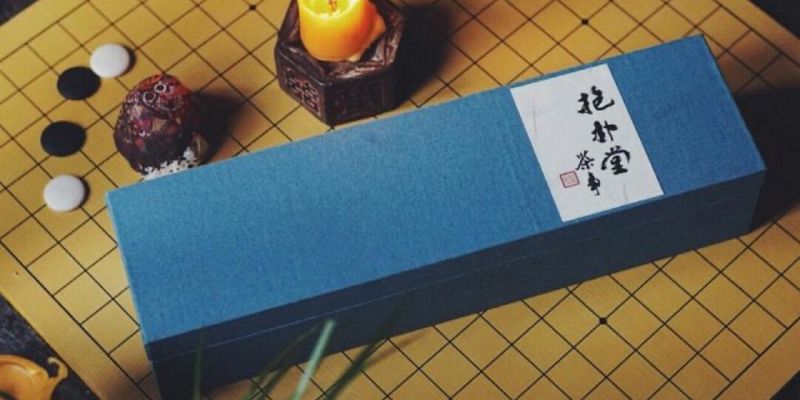 A long blue gift box on a wooden go board, beside black and white stones and a small lit candle