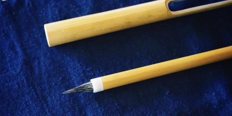 A fine calligraphy brush and its hollow bamboo travel case lying side by side on dark blue fabric