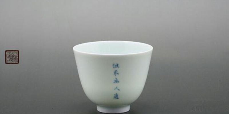A single pale celadon tea cup with vertical blue calligraphy, photographed against a grey studio background