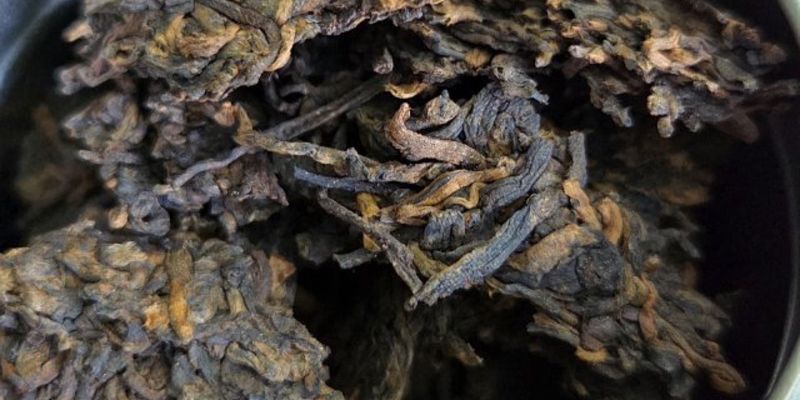 A close-up of dark, broken puer tea leaves inside an open storage jar, with a torn paper wrapper bearing a red seal