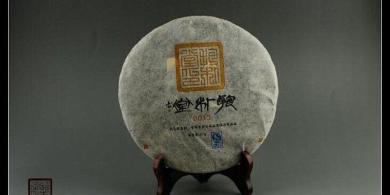 A round raw puer cake wrapped in paper, marked "抱朴堂 0013", displayed on a wooden stand against a grey backdrop